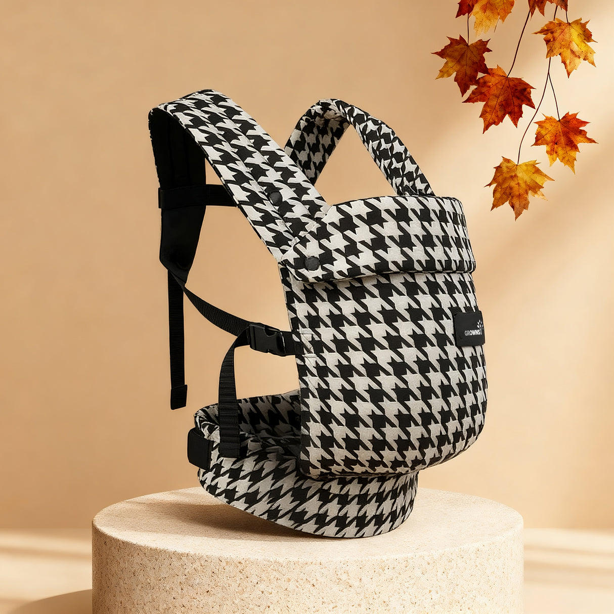 Houndstooth baby carrier with adjustable straps, displayed on a stand with autumn leaves. For product page showcasing design.