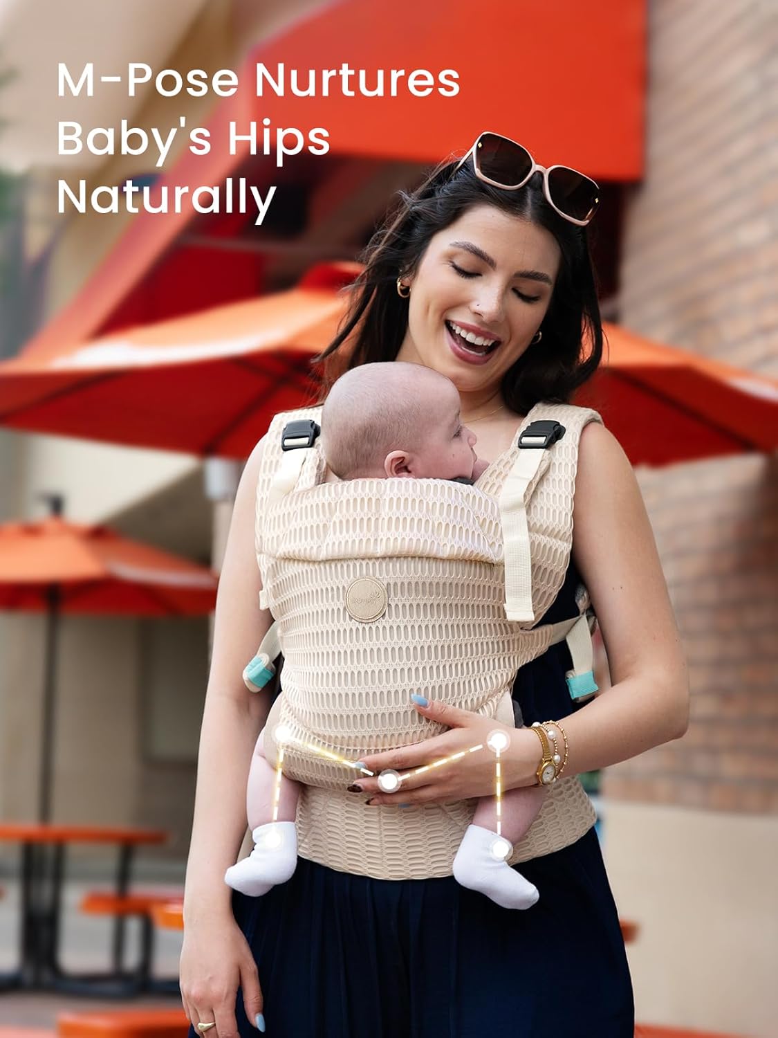 Woman carrying baby in beige mesh carrier demonstrating ergonomic M-pose hip support with graphic overlay.