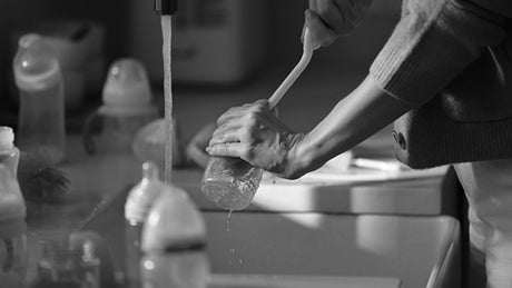 Washing a baby bottle under running water in a kitchen, GROWNSY baby care products visible.