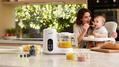 GROWNSY baby food maker on kitchen counter, mom feeding baby in high chair, fresh fruit nearby