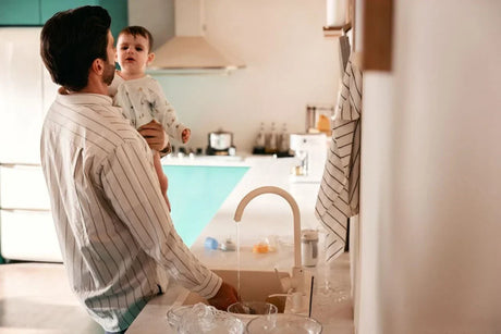 Dad washing baby bottles while holding baby in the kitchen