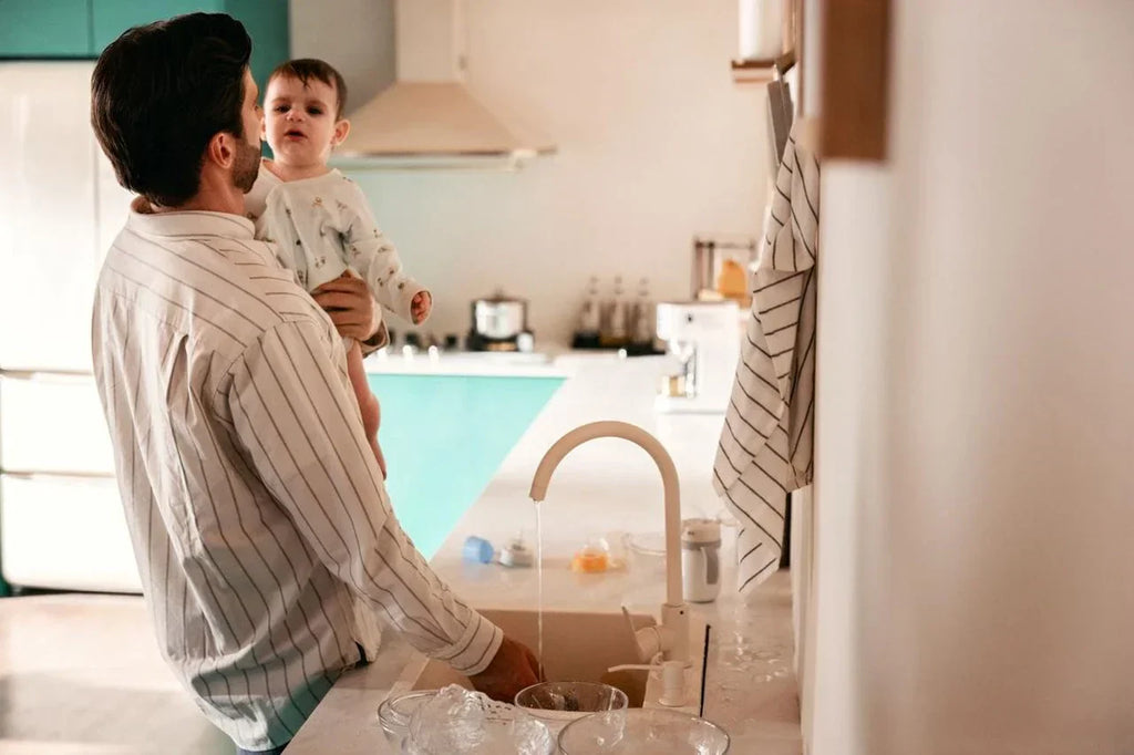 Dad washing baby bottles while holding baby in the kitchen