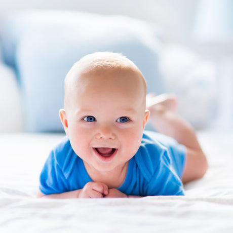 A smiling baby with blue eyes wearing a blue onesie lying on their stomach on white bedding