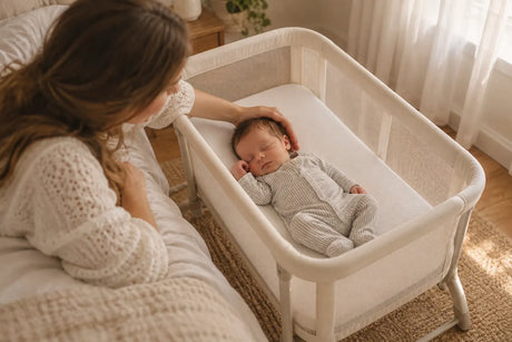 Newborn sleeping in a cream mesh bassinet as their mother gently cradles their head in a softly lit bedroom.