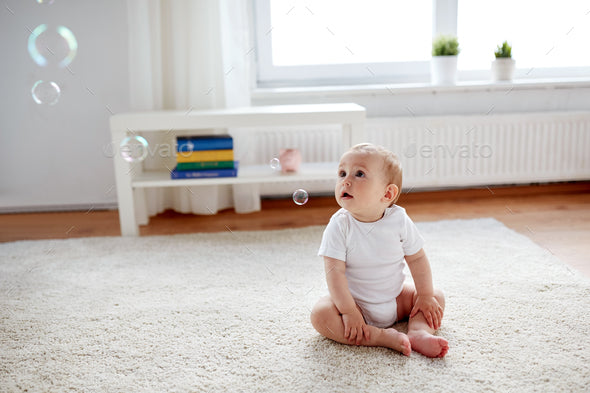 Baby sitting independently on a carpet looking up at floating soap bubbles