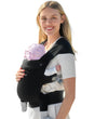 Smiling woman wearing a black ergonomic baby carrier with adjustable straps, comfortably supporting a baby with pink bonnet.